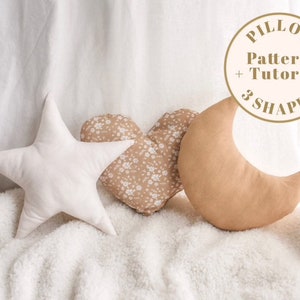 May include: Three plush pillows in the shapes of a star, heart, and crescent moon. The star is white, the heart is brown with a floral pattern, and the moon is tan. The pillows are arranged on a white fluffy surface. A circle with the text "PILLOW Pattern + Tutorial 3 SHAPES" is in the upper right corner of the image.