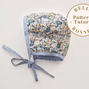 Op de afbeelding: Een blauwe en witte baby muts met bloemen en een blauwe strik. De muts heeft de tekst "Belle Pattern + Tutorial Bonnet" op een witte cirkel.