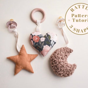 Op de afbeelding: Een DIY-patroon en tutorial voor een babyratel om een ​​ratel te maken met drie vormen: een hart, een ster en een halve maan. De ratel is gemaakt van stof en heeft een houten ring aan de bovenkant.