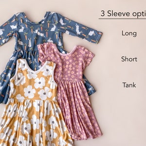 Pode incluir: Tr&ecirc;s vestidos para meninas com diferentes comprimentos de manga: manga comprida, manga curta e sem mangas. O vestido de manga comprida tem um fundo azul com coelhos brancos e cenouras. O vestido de manga curta tem um fundo rosa com flores brancas. O vestido sem mangas tem um fundo amarelo com flores brancas. O texto "3 Sleeve options" est&aacute; no topo da imagem, com "Long", "Short" e "Tank" listados abaixo.