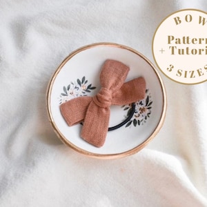 Pode incluir: Um laço de tecido castanho com um elástico preto para cabelo. O laço está sobre um prato branco e dourado com um padrão floral. O texto "BOW Pattern + Tutorial 3 SIZES" está num círculo castanho.