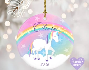 May include: A round ceramic ornament featuring a white unicorn with a multicolored mane and tail, standing on white clouds. A rainbow and stars decorate the background. The text Olivia and 2026 are printed on the front. It hangs from a gold cord.