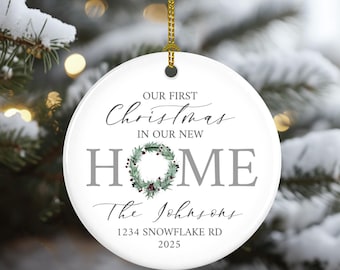 May include: A white ceramic Christmas ornament with gold string. The ornament has the text "OUR FIRST CHRISTMAS IN OUR NEW HOME" and "The Johnsons" with the address "1234 SNOWFLAKE RD" and the year "2025". A wreath design is in the center.