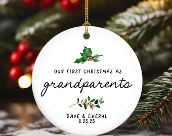Op de afbeelding: Ronde witte kerstbal met een gouden ophangkoord. De tekst luidt "OUR FIRST CHRISTMAS AS grandparents", met de namen "DAVE & CHERYL" en de datum "8.25.25". Versierd met hulst- en bessenillustraties.