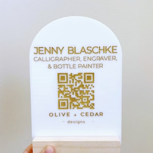 Könnte beinhalten: Ein weißes Acrylschild mit goldfarbenem Schriftzug und einem QR-Code. Auf dem Schild steht "JENNY BLASCHKE, CALLIGRAPHER, ENGRAVER, & BOTTLE PAINTER" und "OLIVE + CEDAR - designs". Das Schild ist auf einem hellen Holzsockel montiert.