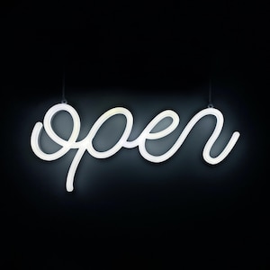 Peut inclure: Enseigne lumineuse blanche en néon avec le mot "open" écrit en cursive. Le signe est suspendu par deux fines chaînes sur un fond noir. L'enseigne lumineuse est un élément décoratif.