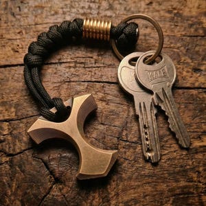 Op de afbeelding: Een bronzen sleutelhouder met een uniek kruisvormig ontwerp. Het heeft een zwart geweven koord, een goudkleurige metalen accent en een metalen ring met twee sleutels. De sleutels zijn zilverkleurig en hebben het woord "KALE" erop.