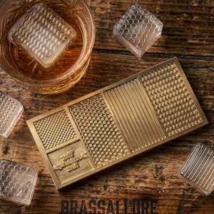 Op de afbeelding: Een messing ijsblokjesvorm met verschillende textuurpatronen, samen met verschillende ijsblokjes met patronen en een glas amberkleurige vloeistof. Het woord "BRASSALLURE" staat onderaan gedrukt. De scène speelt zich af op een houten oppervlak.