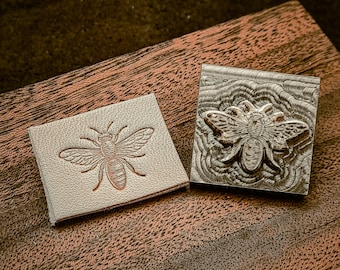 May include: Two square stamps with a bee design. One stamp is a light tan leather square with a detailed bee imprint. The other is a metal stamp with a bee design and a carved background. Both are on a wooden surface.