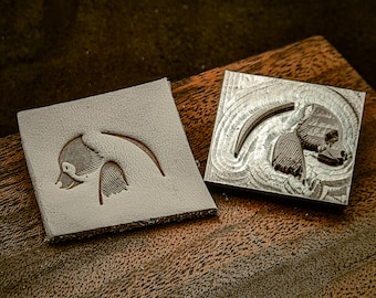 May include: Two square blocks, one gray and one silver, with a bird design. The gray block has a carved bird profile. The silver block is a metal stamp with a detailed, engraved bird image, likely for imprinting.