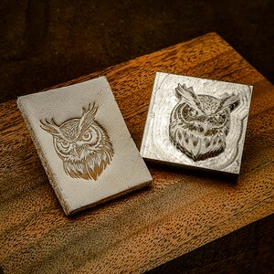 Könnte beinhalten: Zwei Stempel aus Metall mit einem Eulenmotiv. Die Stempel liegen auf einer Holzoberfläche. Das Eulenmotiv ist detailliert und kunstvoll.