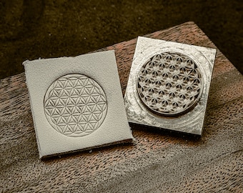 May include: Two metal stamps with a geometric flower of life design. The stamps are on a wooden surface.
