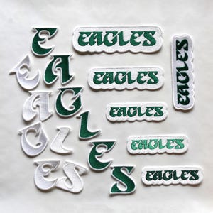 May include: A collection of embroidered patches featuring the word "EAGLES" in various styles and sizes. The patches have a white background with green lettering and border. Some patches feature individual letters.