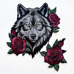 May include: Embroidered iron-on patch featuring a wolf's head with a fierce expression, surrounded by burgundy roses with green leaves.