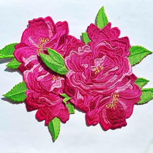 May include: A cluster of pink embroidered flowers with green leaves. The flowers are arranged in a bouquet-like shape.