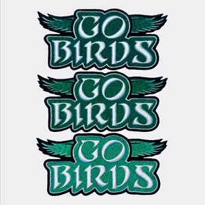 May include: Three embroidered patches with the text "GO BIRDS" in white, outlined in teal, and set against a teal background. Each patch features a pair of green wings above the text, with a black border.