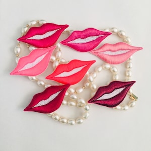 May include: A string of white pearls with six embroidered lips in various shades of pink and red.