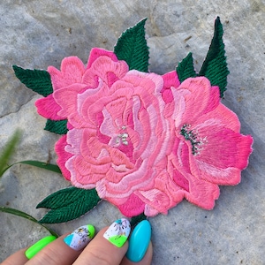 May include: A pink embroidered flower patch with green leaves. The patch is made of fabric and has a textured surface.