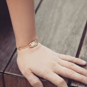May include: A delicate gold chain bracelet with a rectangular, light pink gemstone pendant. The bracelet is on a wooden surface.