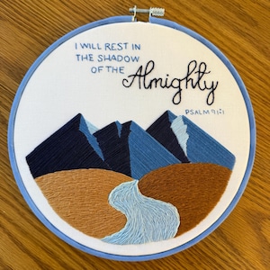 May include: Embroidered hoop art with a blue, white, and brown mountain landscape and a flowing river. The text reads "I will rest in the shadow of the Almighty, Psalm 91:1".