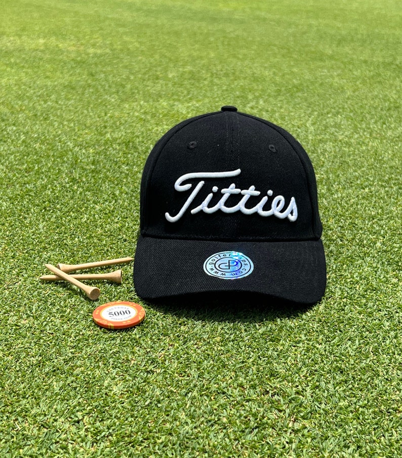 May include: Black baseball cap with a white embroidered "Titties" logo. The cap has a black adjustable strap and a small black and white logo on the back.