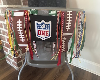 May include: A high chair banner with a football theme. The banner features brown football shapes, the NFL logo with the word "ONE", and colorful ribbons. The banner is designed for a first birthday celebration.