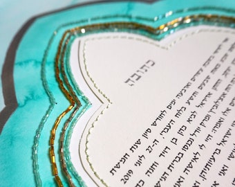 Op de afbeelding: Een witte ketubah met Hebreeuwse tekst, omringd door een turquoise rand met gouden en zilveren kralen. De tekst luidt "Ketubah" en bevat de datum 27 juni 2019.