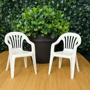 May include: Two small, white plastic chairs with armrests are displayed on a wooden surface. The chairs are in front of a dark pot with a green plant. The background is a green hedge. The chairs are miniature and have a classic design.