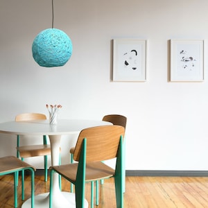 Puede incluir: Una mesa redonda de comedor blanca con cuatro sillas. Dos sillas son de color verde azulado con asientos de madera y dos sillas son de madera natural con asientos de madera. Una lámpara de bola de hilo azul cuelga del techo sobre la mesa.
