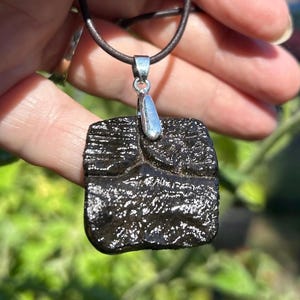 May include: A dark, textured pendant with a silver bail hangs from a brown leather cord. The pendant is square-shaped and appears to be made of a dark, possibly natural, material. The background is blurred green foliage.