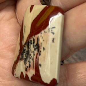 May include: A silver chain necklace with a rectangular pendant made of polished, multi-colored stone. The stone has a white base with brown and red swirls and black speckles.