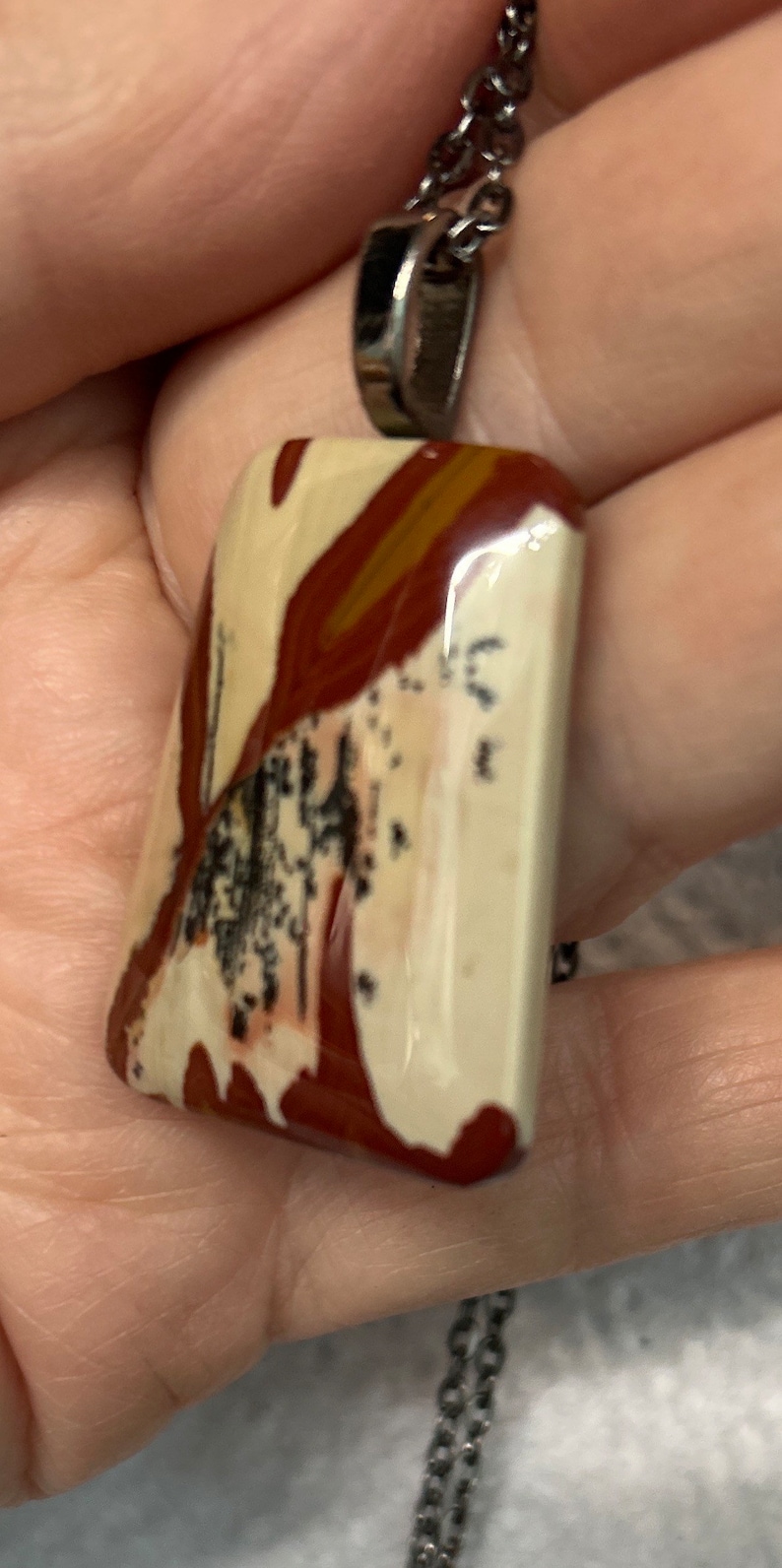 May include: A silver chain necklace with a rectangular pendant made of polished, multi-colored stone. The stone has a white base with brown and red swirls and black speckles.
