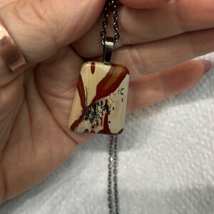 May include: A silver chain necklace with a rectangular pendant made of a colorful, patterned stone. The stone has shades of red, white, and black.