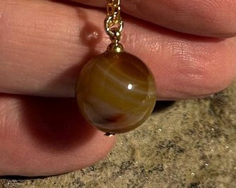 May include: A spherical brown and tan banded agate gemstone pendant suspended from a delicate gold-toned metal chain. The stone features natural swirling patterns with translucent layers and a polished, glossy surface finish reflecting light.