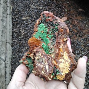 May include: A rough, brown rock with green and yellow mineral deposits. The rock is being held in a hand.