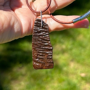 May include: A brown, textured, rectangular pendant hangs from a brown leather cord. The pendant is attached to the cord with a silver jump ring.