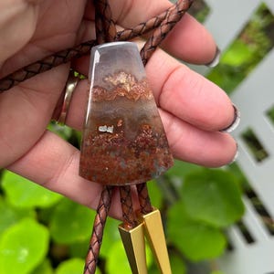 May include: A bolo tie featuring a polished, trapezoid-shaped stone pendant with brown, red, and gray patterns. The pendant is suspended from a braided brown leather cord, with gold-colored metal tips.