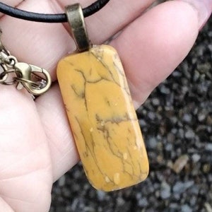 May include: A yellow and brown marbled stone pendant on a black leather cord necklace. The pendant is rectangular in shape and has a bronze-colored metal bail.