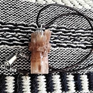 May include: A brown crystal pendant with a silver setting hangs from a brown leather cord. The pendant is on a black and white patterned surface.