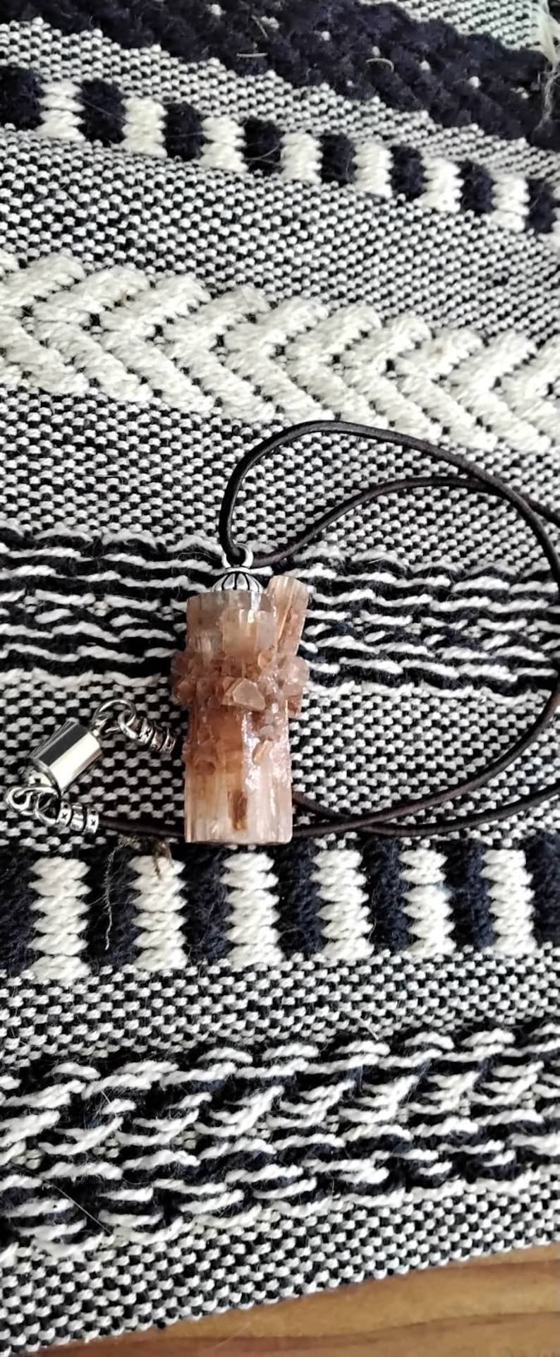 May include: A brown crystal pendant with a silver setting hangs from a brown leather cord. The pendant is on a black and white patterned surface.