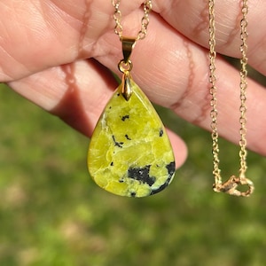 May include: A gold-colored chain necklace with a teardrop-shaped pendant. The pendant is a vibrant yellow-green stone with black inclusions. The necklace is displayed against a blurred green background.