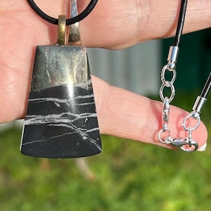 May include: A black and white stone pendant with a silver and gold-toned bail, suspended from a black leather cord. The pendant features a polished, trapezoid shape with a marbled pattern. The cord is secured with a silver clasp.