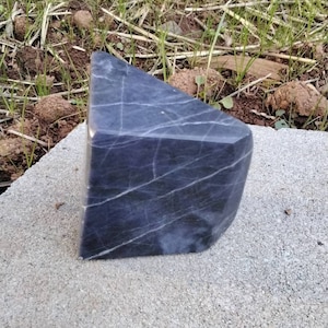 May include: A dark gray, irregularly shaped, polished stone with white veins. The stone is sitting on a gray concrete surface.