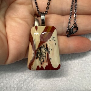 May include: A silver chain necklace with a rectangular pendant featuring a colorful, abstract pattern in shades of red, white, and black.