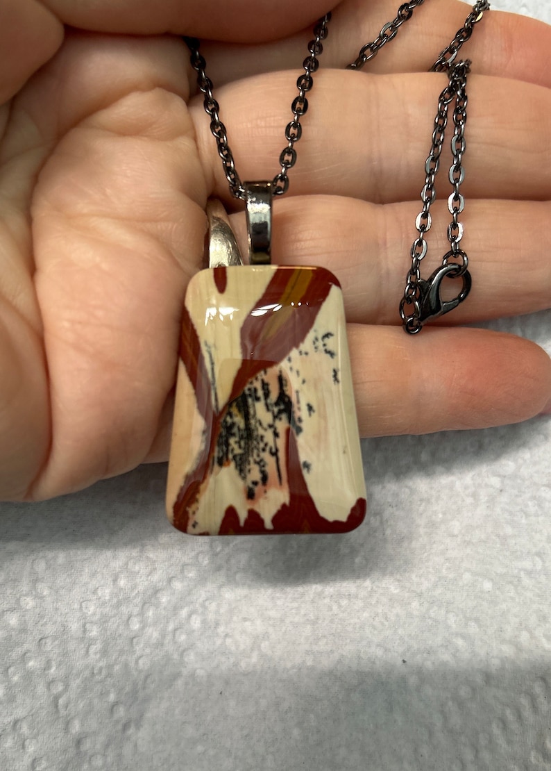 May include: A silver chain necklace with a rectangular pendant featuring a colorful, abstract pattern in shades of red, white, and black.