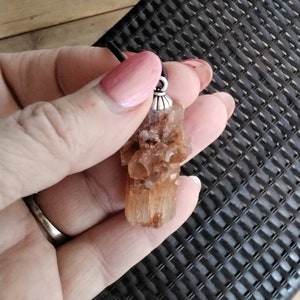 May include: A brown and orange crystal pendant with a silver bail. The pendant is hanging from a black cord.