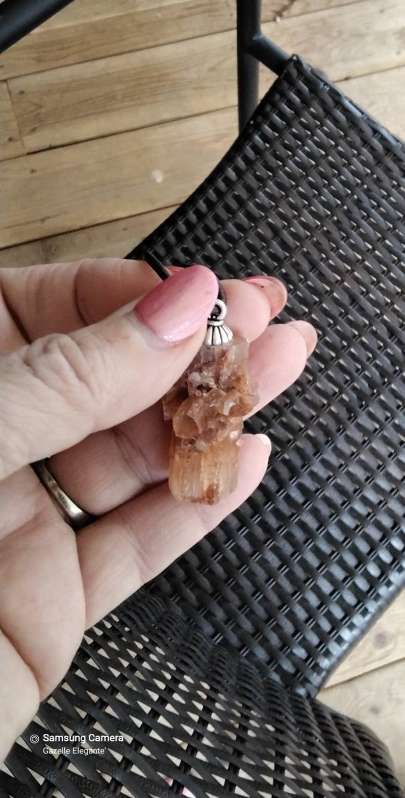 May include: A brown and orange crystal pendant with a silver bail. The pendant is hanging from a black cord.