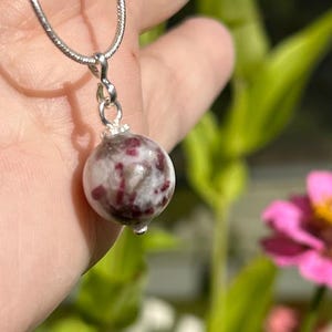 May include: A silver necklace with a round pendant. The pendant is a white and maroon speckled stone. The necklace is held in a hand with a blurred green and pink floral background.