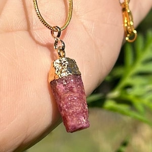 May include: A gold chain necklace with a pink ruby pendant. The ruby is a raw, uncut crystal with a gold-plated top.