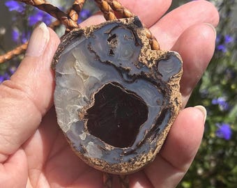 May include: A bolo tie featuring a large, irregular, polished agate stone pendant with a dark brown center, translucent gray banding, and a rough, textured tan outer edge. The pendant is attached to a braided tan leather cord with metal tips.